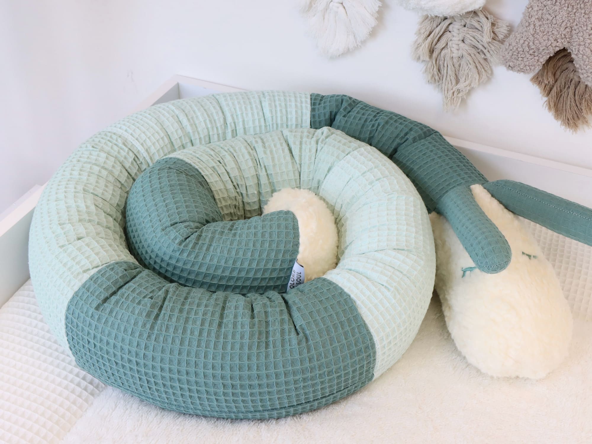 Waffelpique-Puckschnecke in Grün- und Minttönen, aufgerichtet stehend im Babybett, mit flauschigem Kopf aus Teddyplüsch.