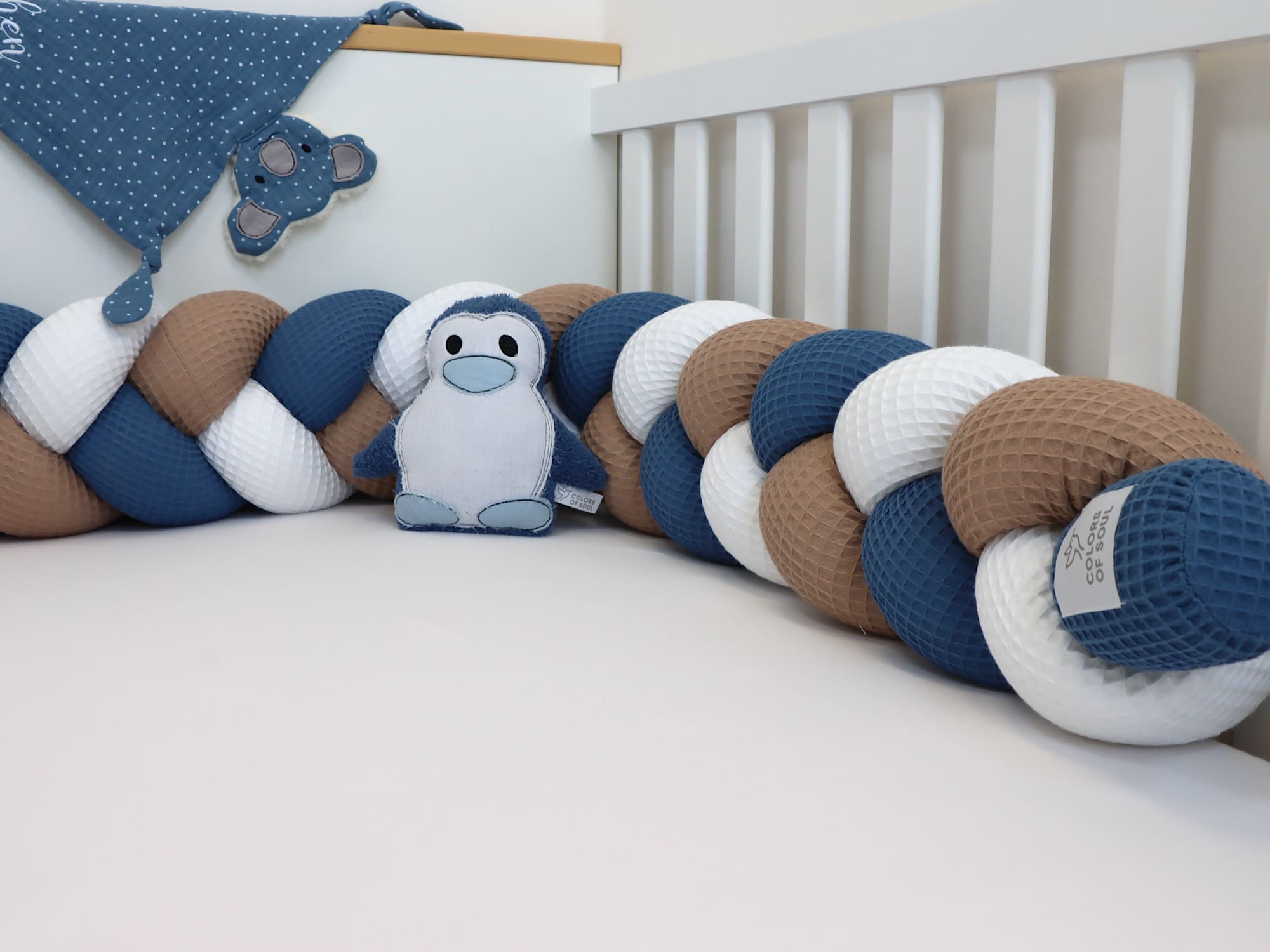 Bettschlange Weiß–Dunkelbeige–Jeansblau aus Waffelpique im Babybett, dekoriert mit Pinguin-Stofftier und blauem Tuch.