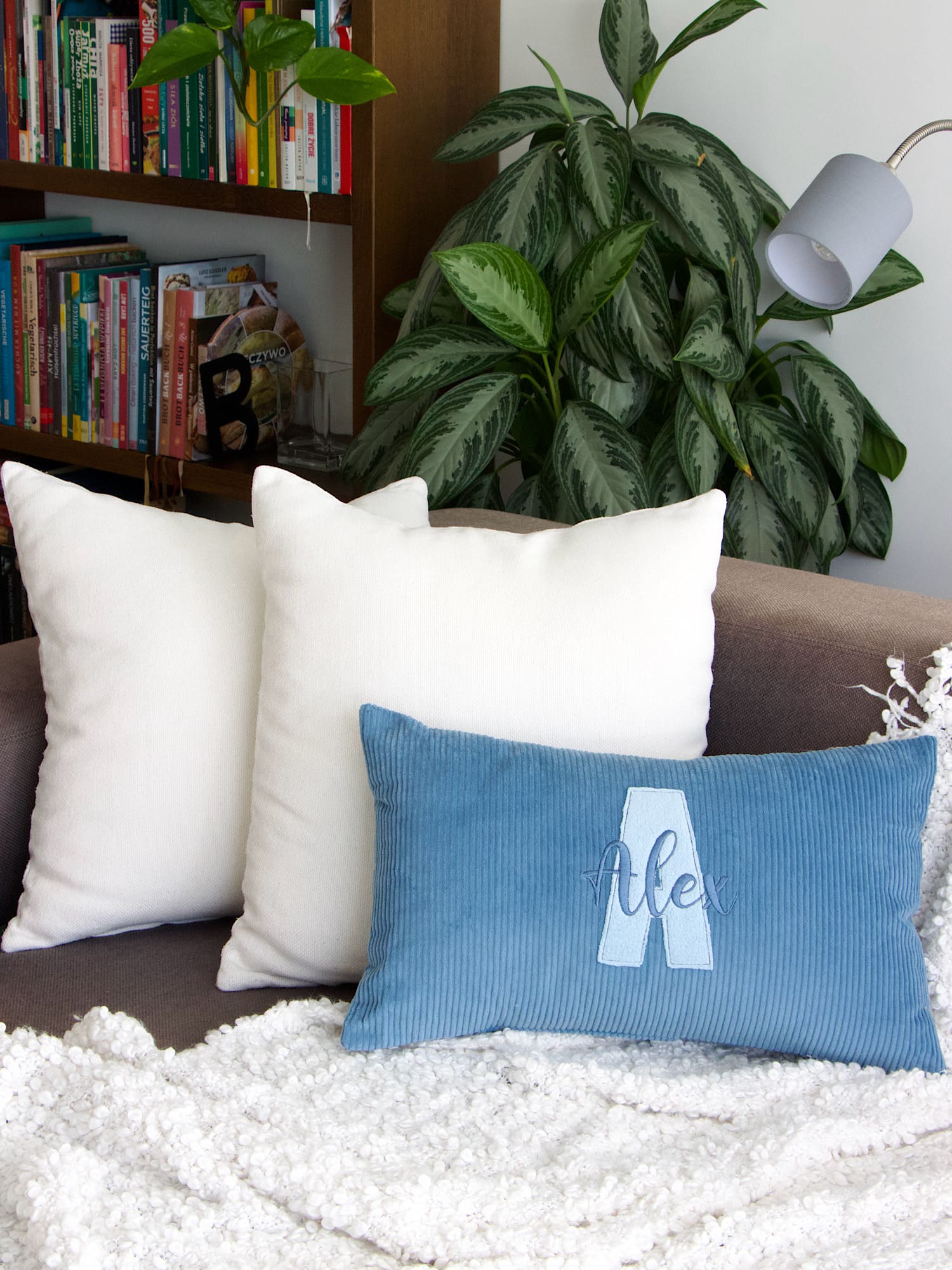 Wohnzimmer-Szene mit personalisiertem Cordkissen in Blau, Bücherregal und grüner Pflanze im Hintergrund