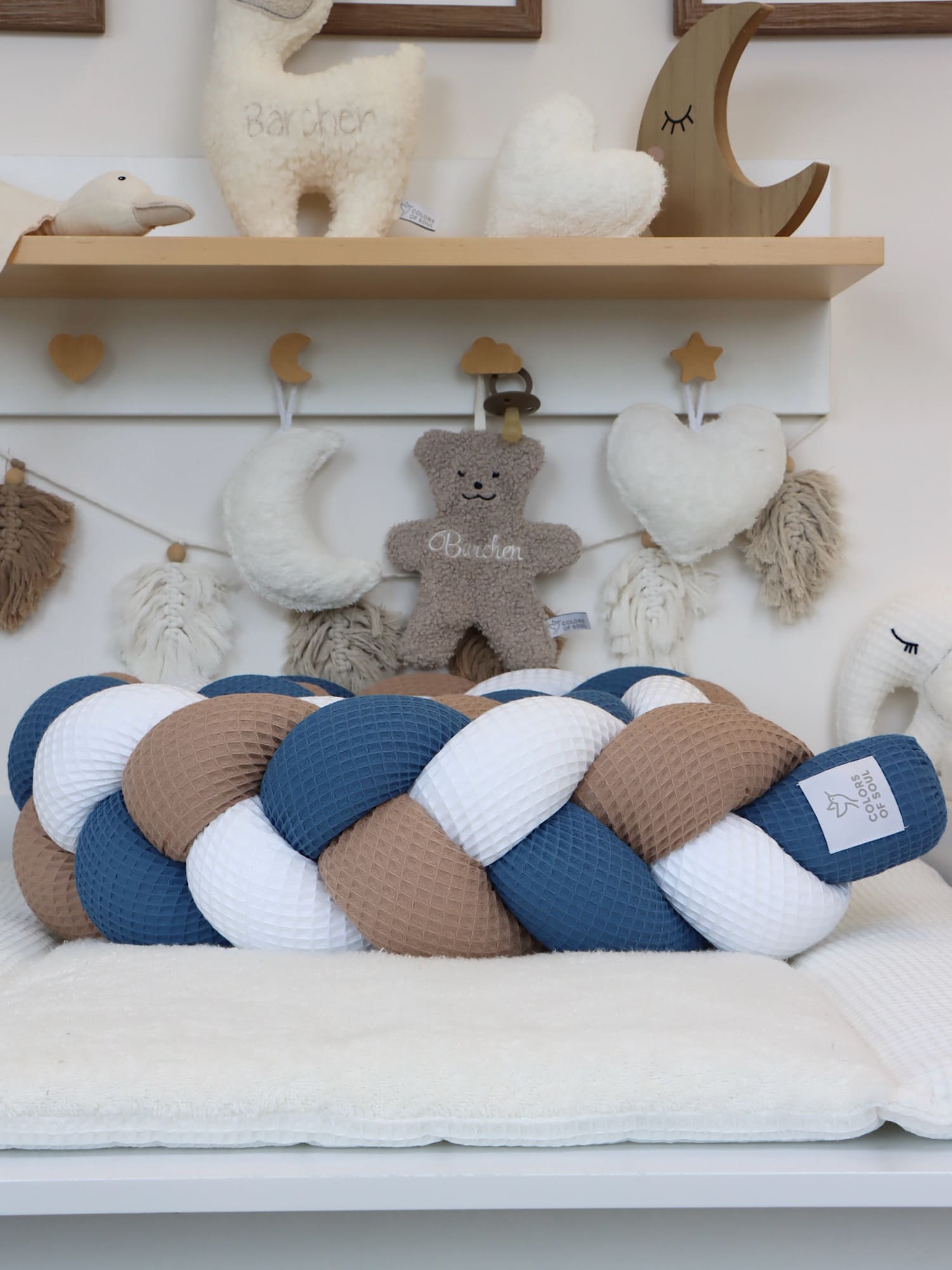 Geflochtene Bettschlange in zeitloser Farbkombination Weiß, Dunkelbeige & Jeansblau auf Babybett-Auflage mit Deko im Hintergrund.