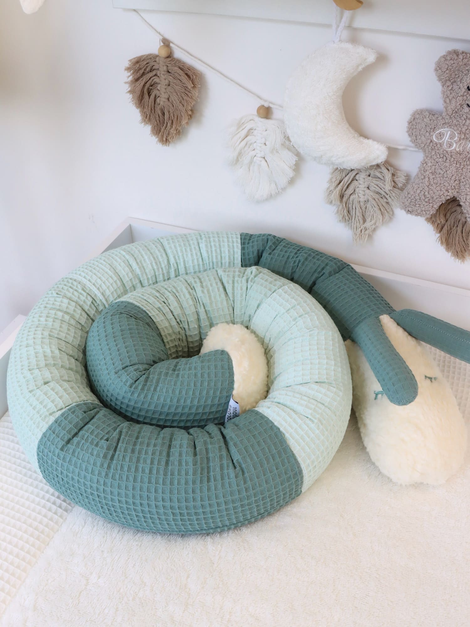 Puckschnecke in Altgrün und Pastellmint, spiralförmig aufgestellt im weißen Babybett mit Naturdeko im Hintergrund.