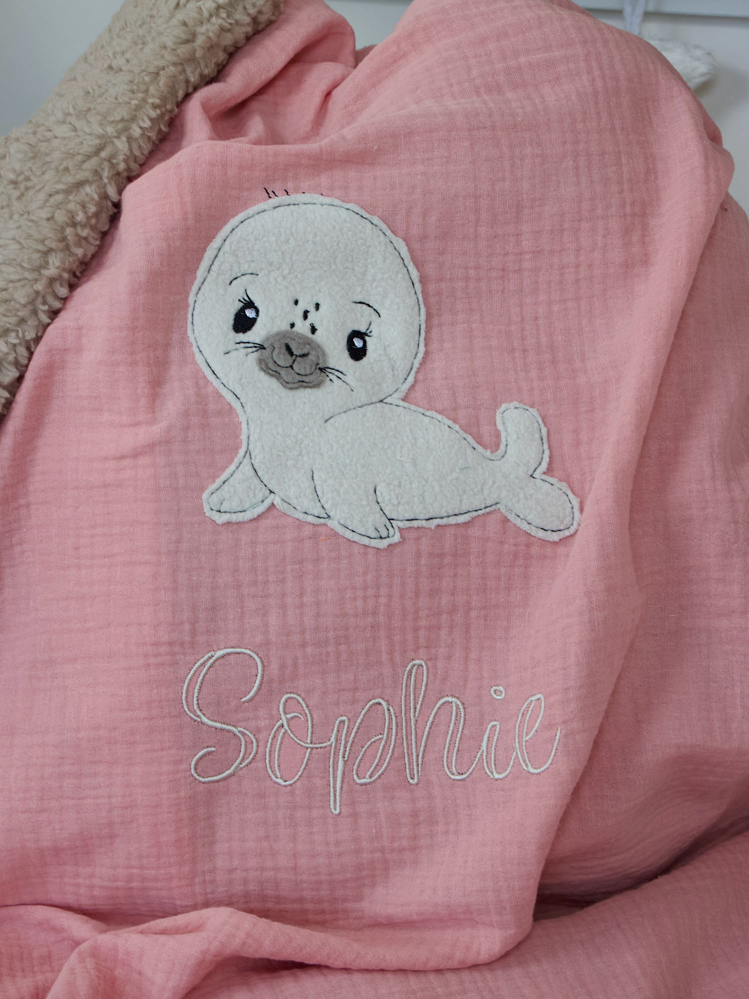 Nahaufnahme des gestickten Seehund-Motivs auf der Musselin-Babydecke in Lachsrosa mit sandfarbenem Kuschelplüsch.