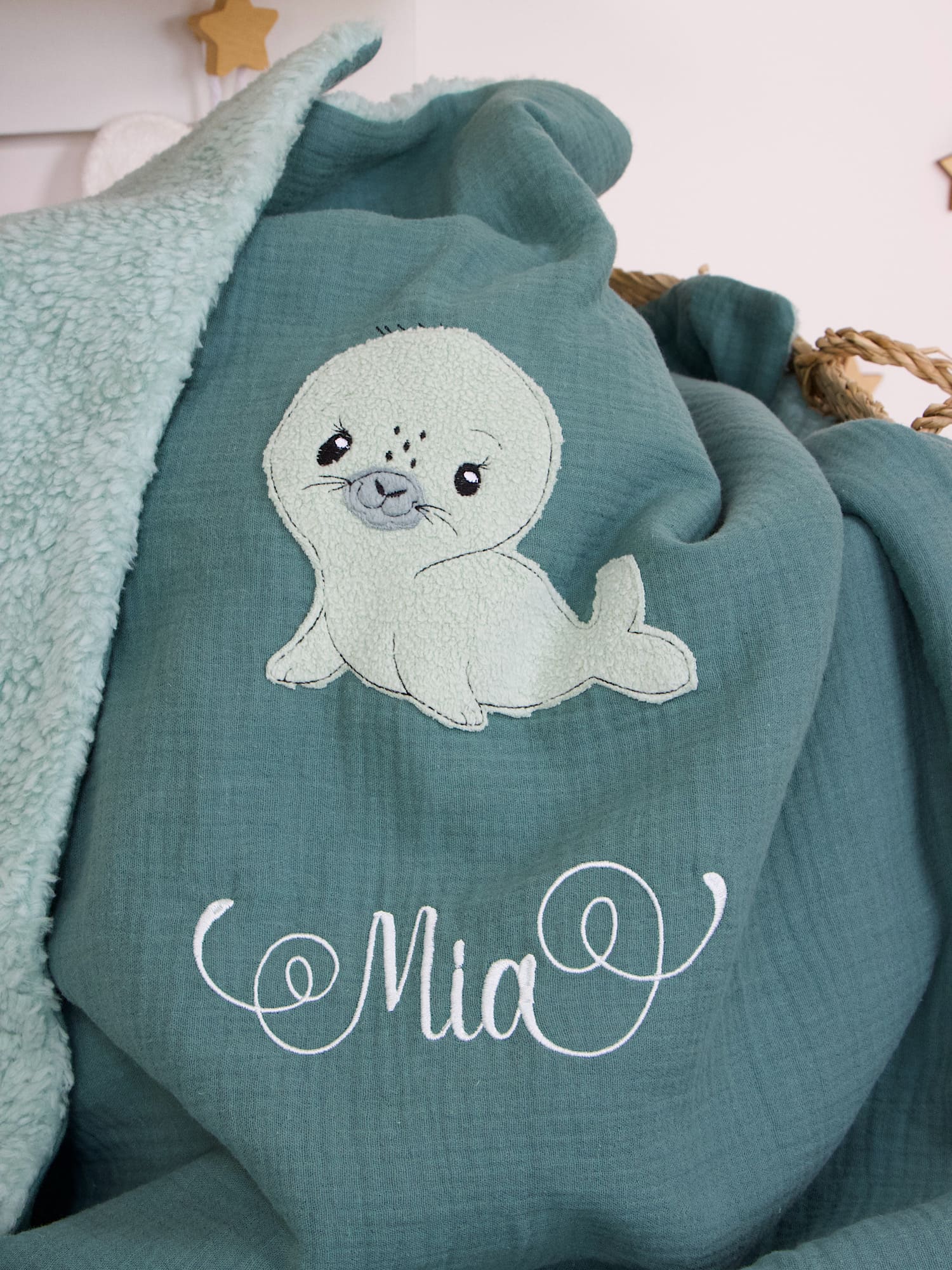 Nahaufnahme der bestickten Babynamen-Stickerei "Mia" in Weiß auf Musselin Dunkelmint mit Seehund-Motiv