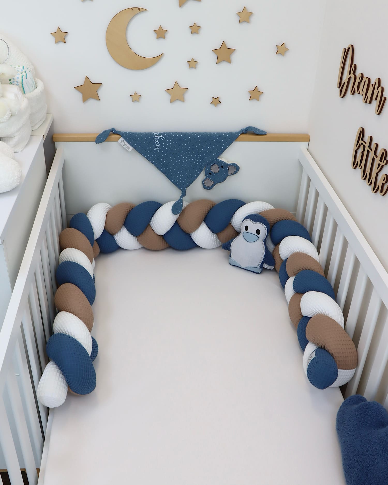 Babybett mit geflochtener Bettschlange in Weiß, Dunkelbeige und Jeansblau, dekoriert mit Sternen-Wanddeko und blauem Tuch.
