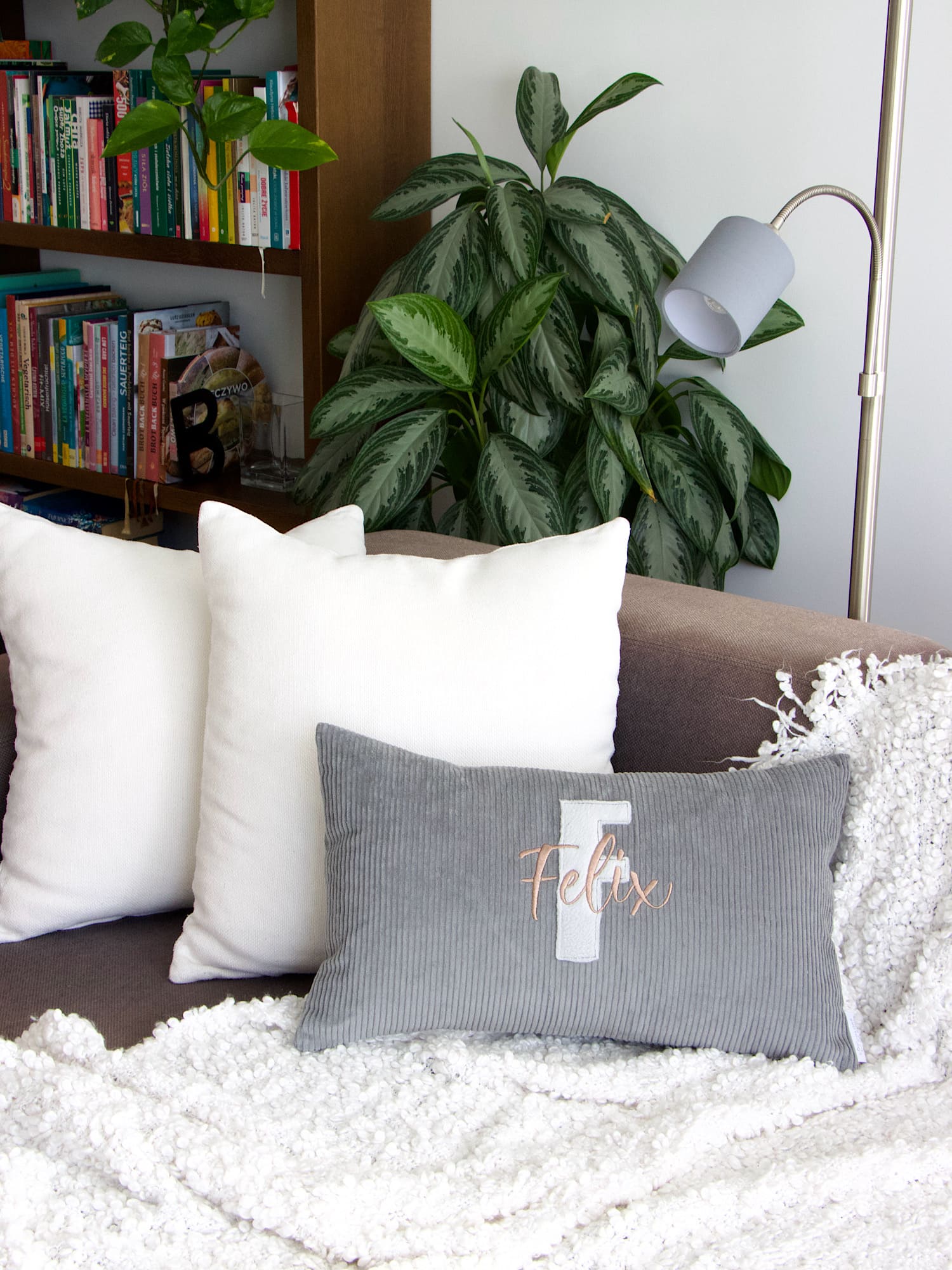 Modernes Wohnzimmer mit personalisiertem Cordkissen in Grau, Bücherregal und Stehlampe im Hintergrund