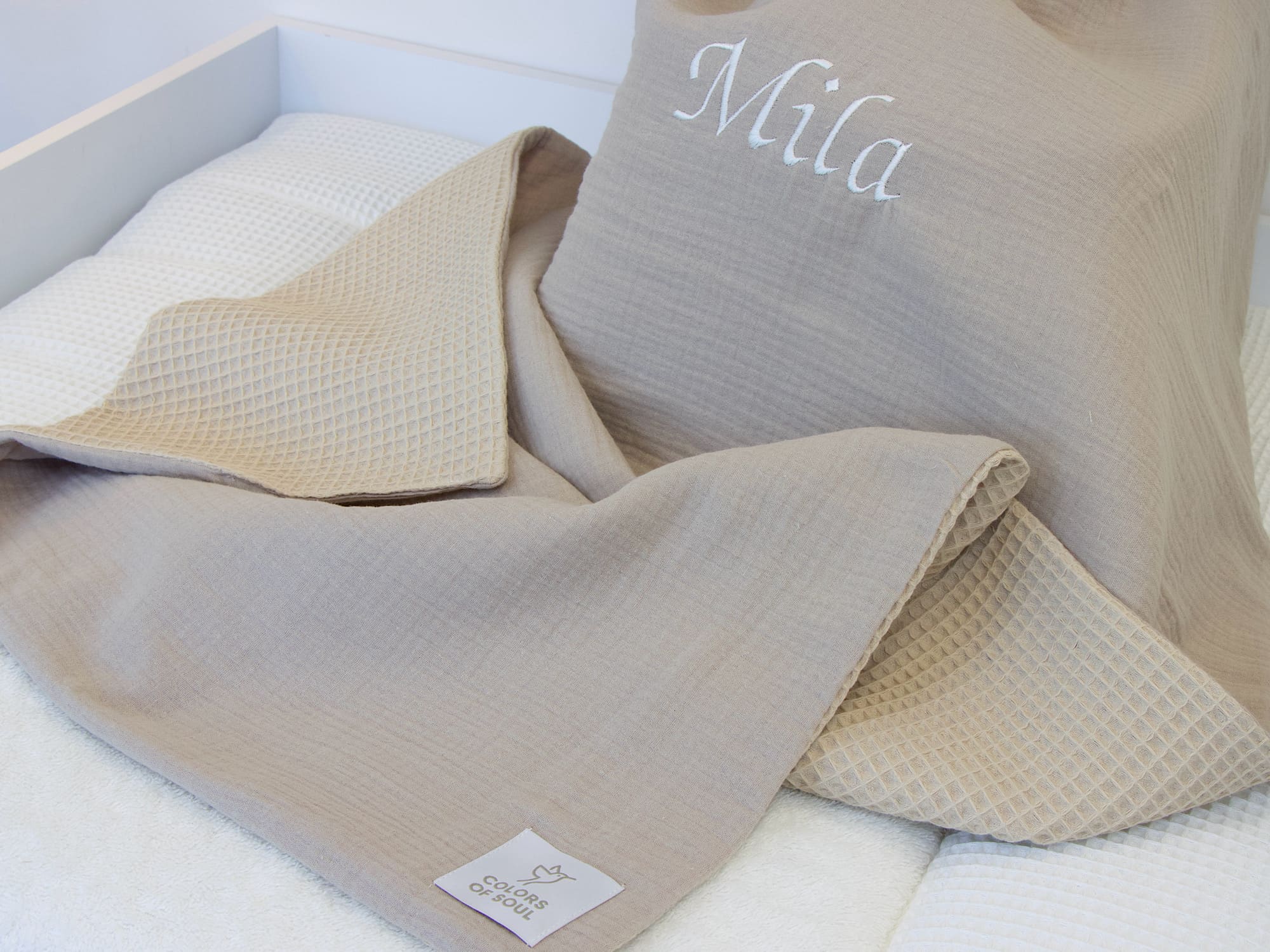 Handgefertigte Babydecke Musselin Beige & Waffelpique Beige mit weißer Namensstickerei, eingerahmt von Babydetails im Kinderzimmer.
