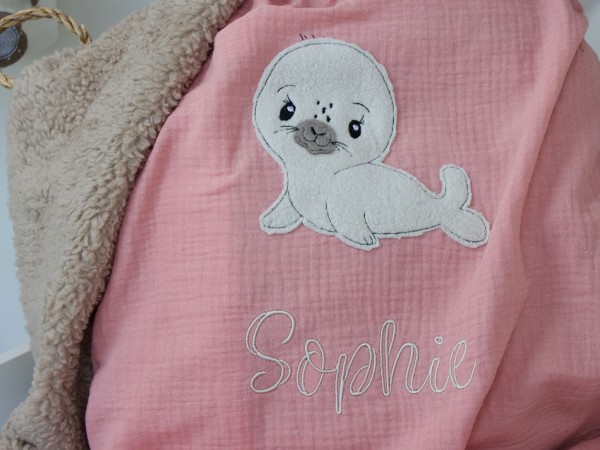 Handgemachte Kuscheldecke aus Musselin Lachsrosa und Teddyplüsch Sand, Seehund-Motiv und Namensstickerei "Sophie" im Babybett.